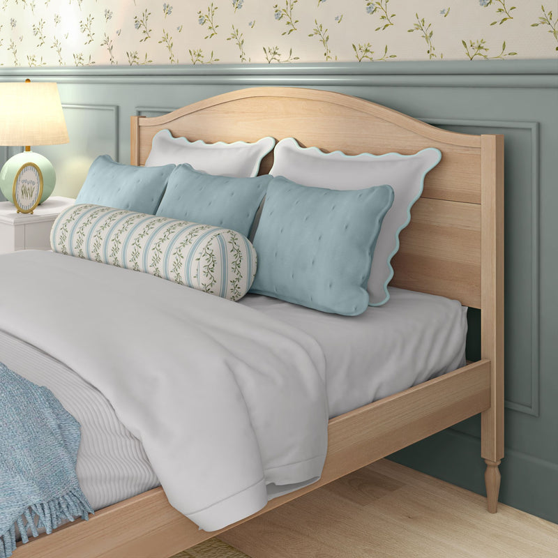 Cozy pastel bedroom interior: light wood headboard, scalloped white-and-blue pillows, striped floral bolster, blue throw blanket and bedside lamp