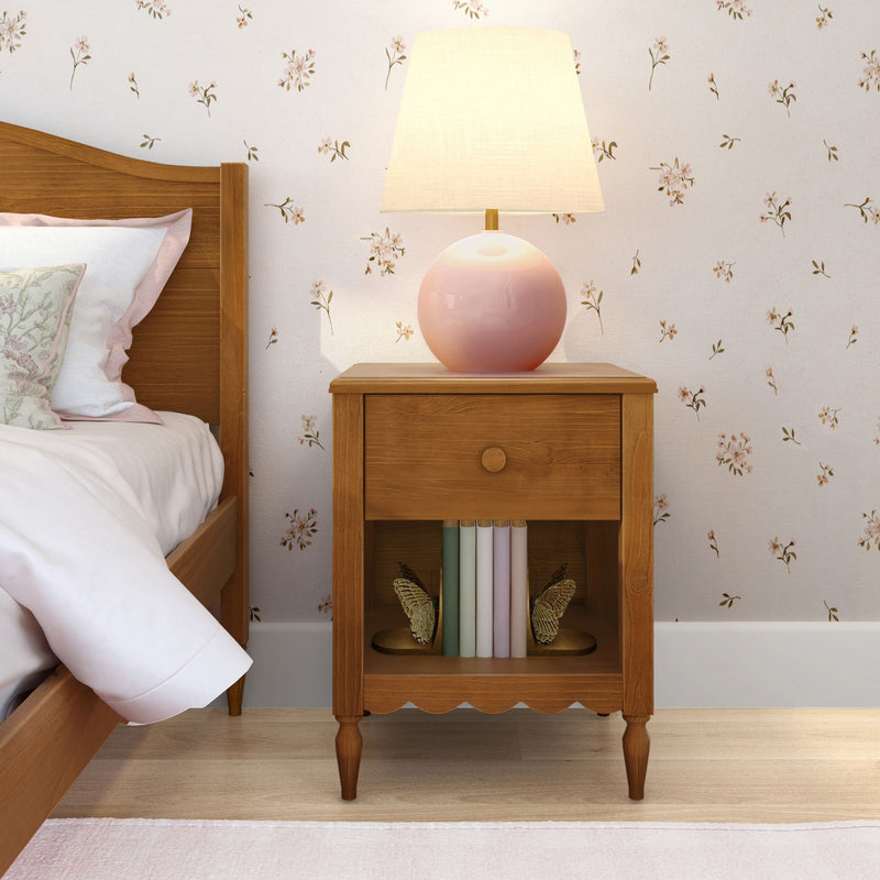 Wooden bedside table with pink ceramic lamp, pastel books and butterfly bookends against floral wallpaper in a cozy cottage bedroom