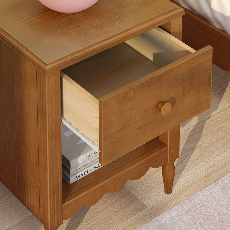 Light brown wooden nightstand bedside table with open drawer and lower shelf holding stacked books, scalloped apron and turned legs on hardwood floor