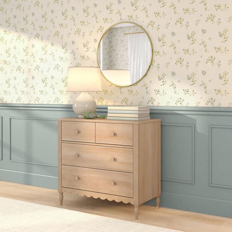 Wooden chest of drawers in a cozy modern farmhouse bedroom with a round gold mirror, table lamp, stacked books and green headphones against pale blue wainscoting and vintage floral wallpaper