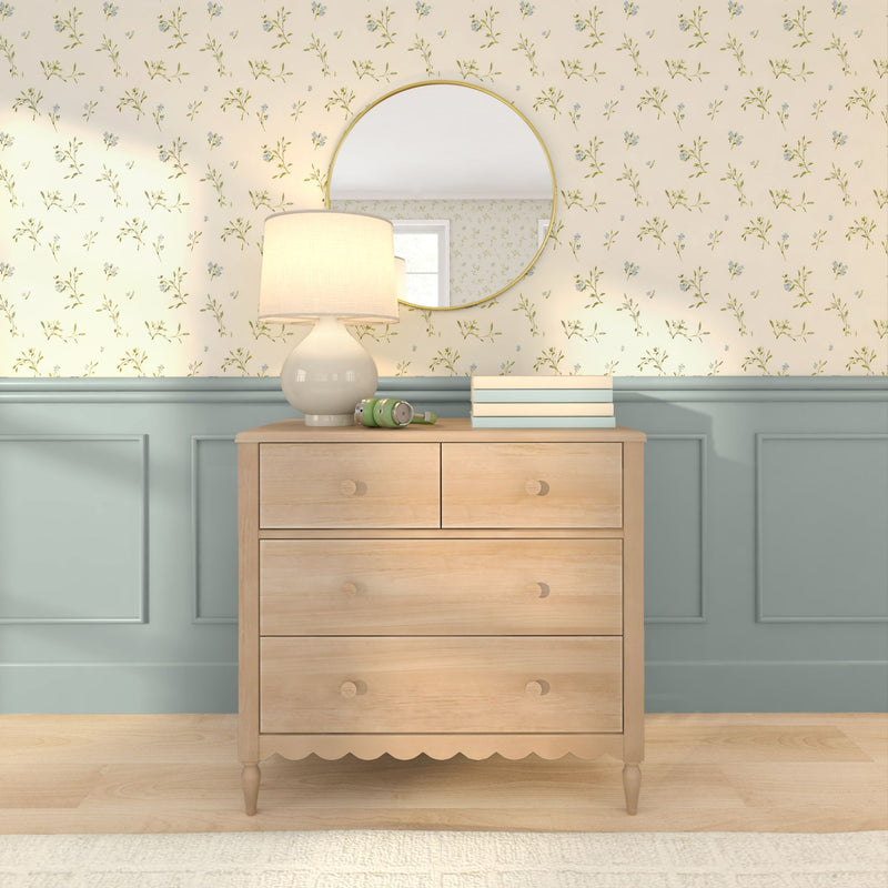 Light wood dresser with ceramic lamp, round gold mirror, stacked books and green headphones against cream floral wallpaper and blue wainscoting — cozy bedroom decor