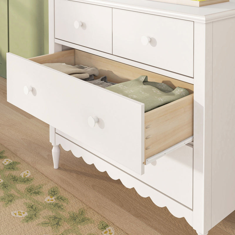 White wooden bedroom dresser with open drawer revealing folded clothes, round white knobs, scalloped base trim and light hardwood floor