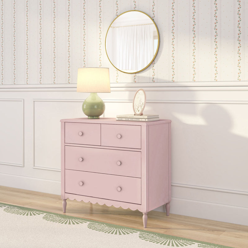 Pink scalloped dresser with round gold mirror, green ceramic lamp and framed print against floral striped wallpaper in a nursery