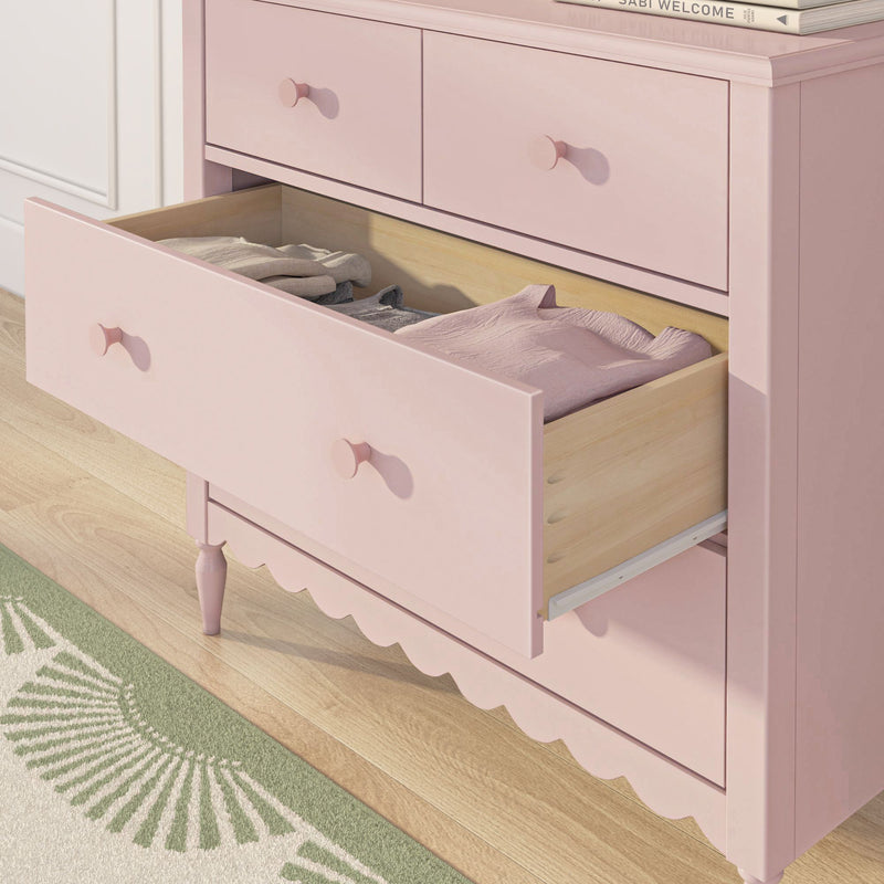 Pink nursery dresser with open drawer of folded baby clothes, round knobs and scalloped trim on light hardwood floor next to green patterned rug