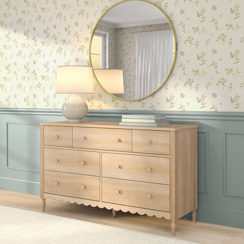 Light wood dresser with beige table lamp and stacked books, round gold mirror reflecting window, floral wallpaper and pastel blue wainscoting, cozy bedroom decor