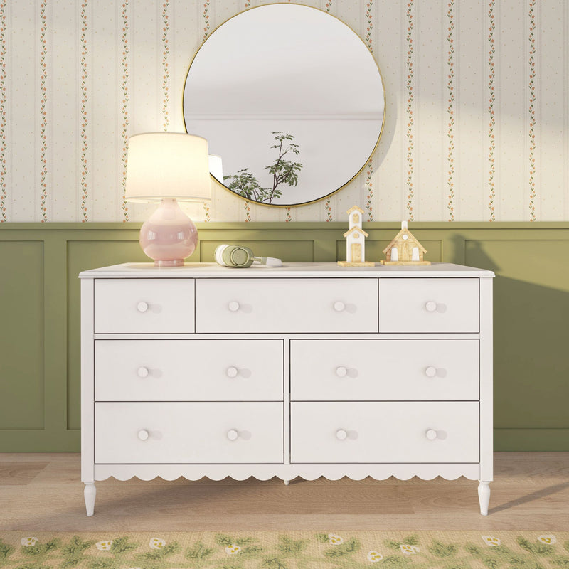 White nursery dresser with round gold mirror, pink lamp, floral wallpaper, green wainscoting and wooden house decor