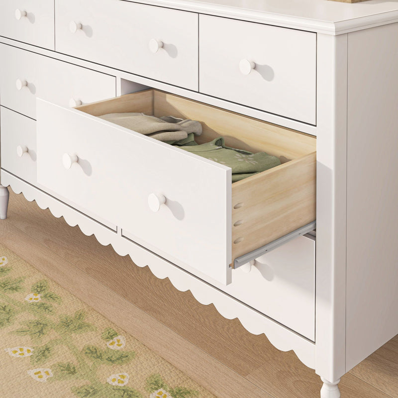 White nursery dresser with open drawer revealing folded baby clothes and natural wood drawer interior, scalloped skirt detail and hardwood floor, children's storage dresser