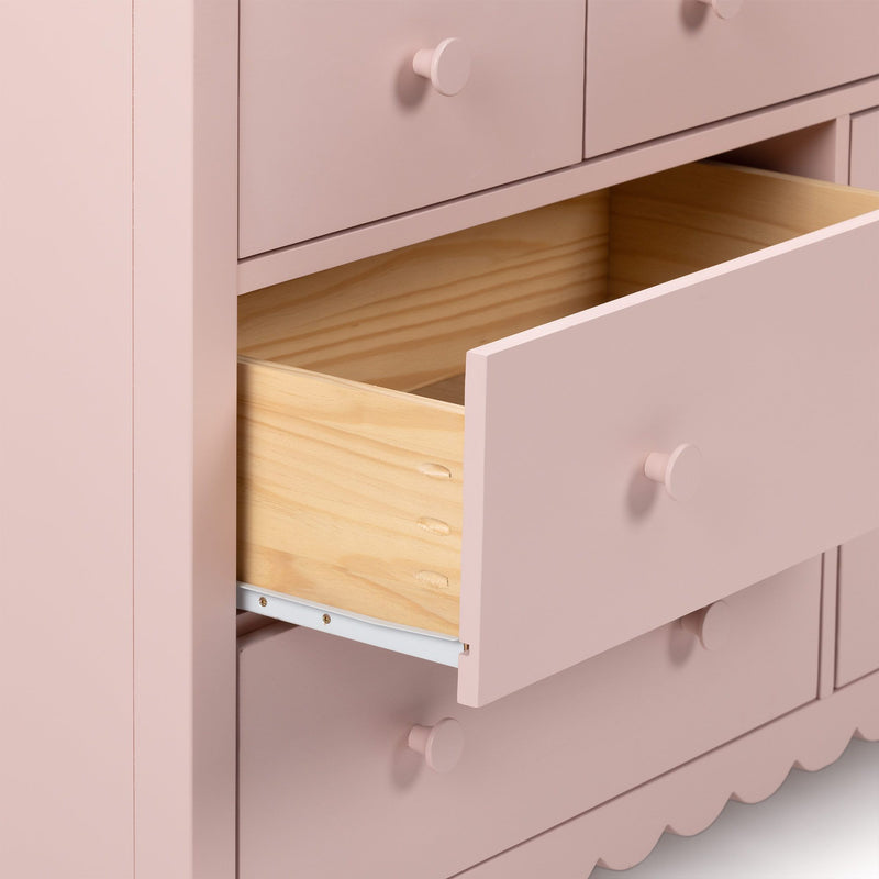 Pastel pink wooden dresser with an open drawer revealing natural pine interior, round knob pulls and scalloped base — stylish nursery or girls' bedroom storage chest