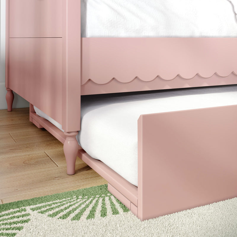 Pink scalloped bed frame with white mattress and under-bed trundle on hardwood floor with green area rug