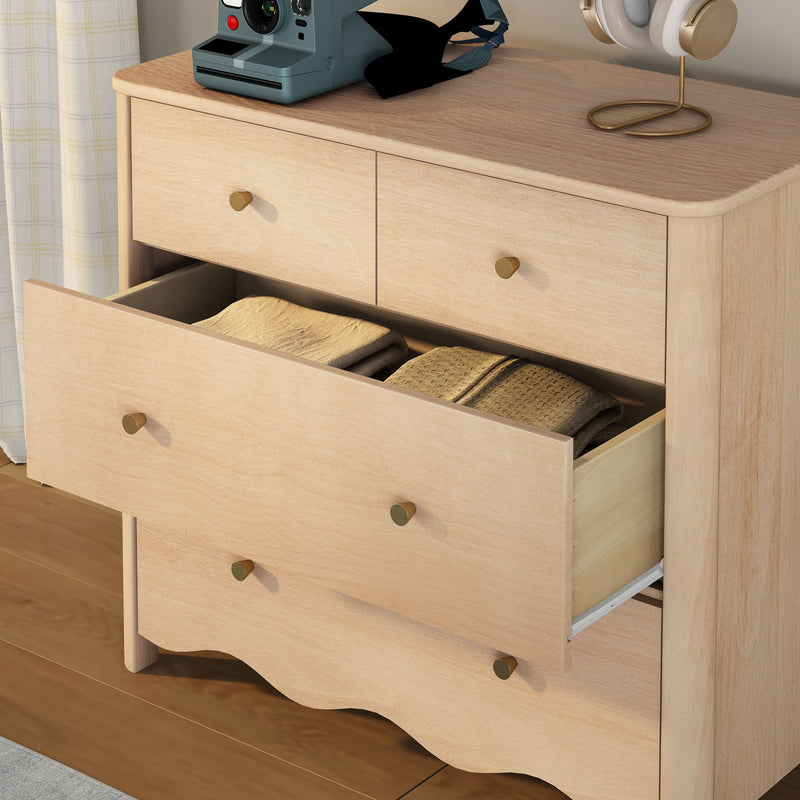 Light wood dresser with open drawer revealing folded blankets, brass knobs, Polaroid camera and gold headphones on top, bedroom storage on hardwood floor
