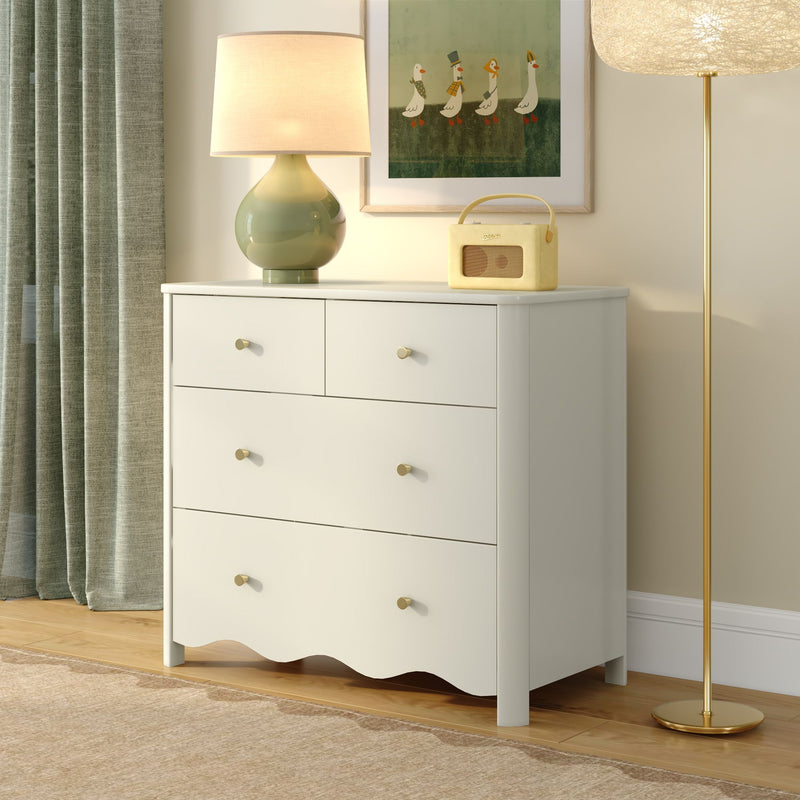 Minimalist white dresser with brass knobs topped by a green ceramic table lamp and retro yellow radio, framed duck artwork and gold floor lamp beside textured green curtains in a cozy modern bedroom interior.