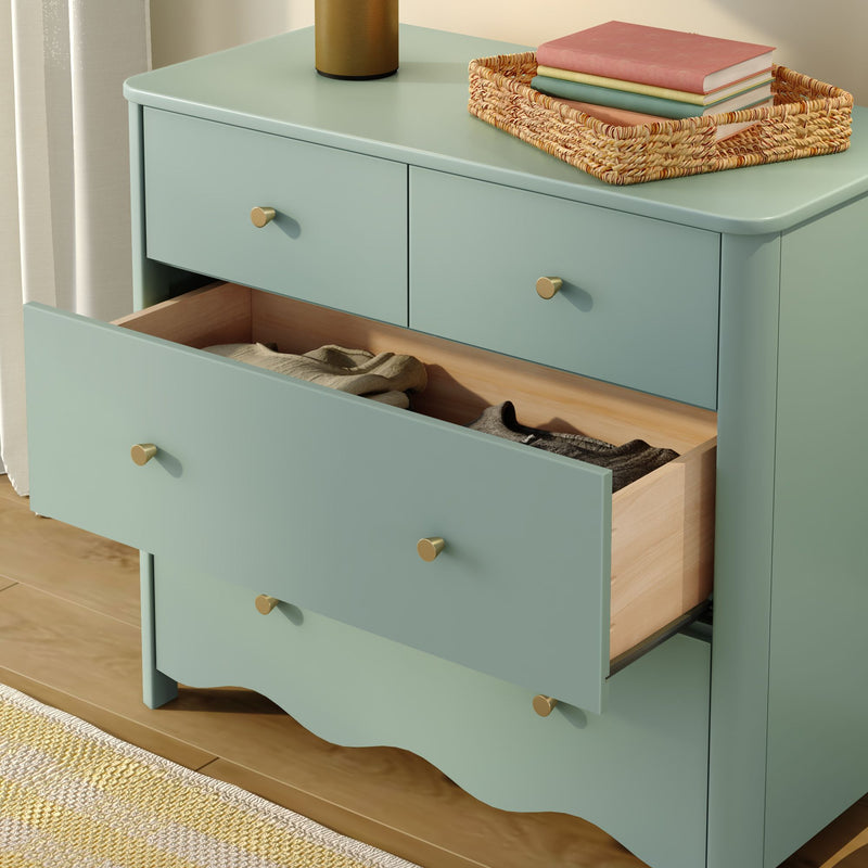 Mint green wooden dresser with open drawer, brass knobs and wicker tray holding stacked books on top — bedroom storage on hardwood floor