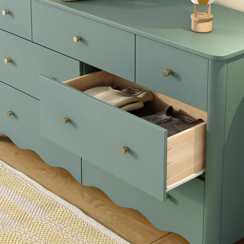 Seafoam green wooden dresser with an open drawer revealing folded sweaters, brass knobs, scalloped base and light hardwood floor