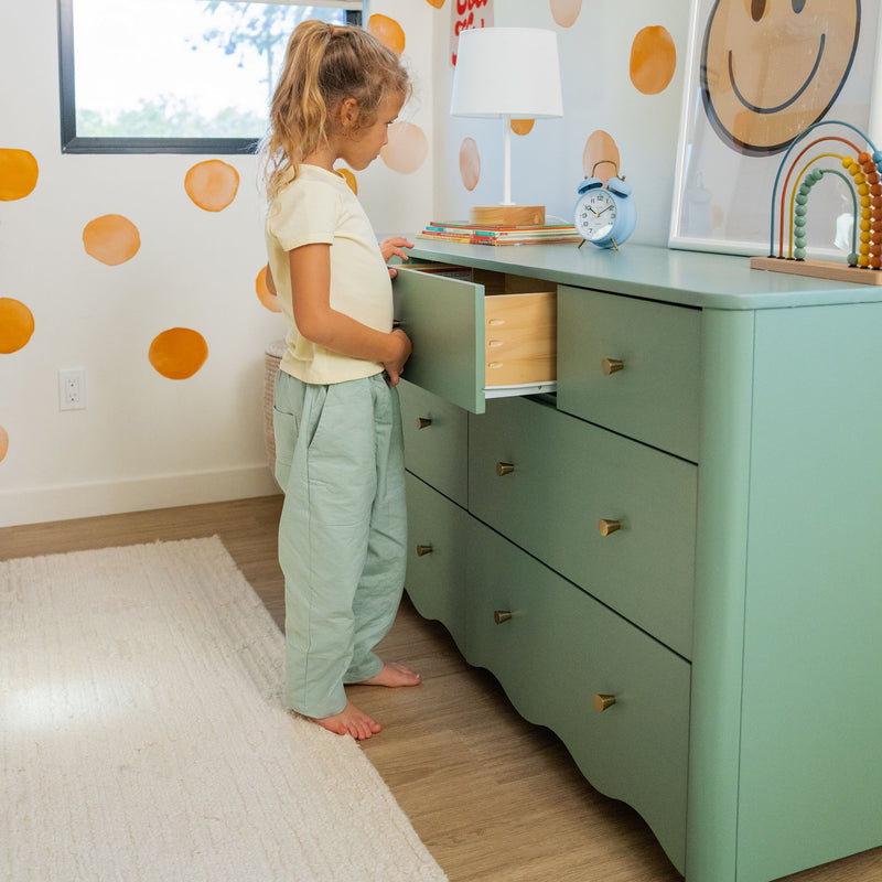 Child opening drawer of mint green dresser in colorful kids' bedroom with orange polka-dot wall, lamp and blue alarm clock