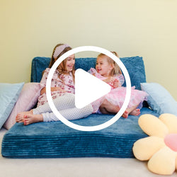 Video of two young girls sitting and laughing on a blue couch, surrounded by colorful pillows and a plush flower, highlighting playful home decor.