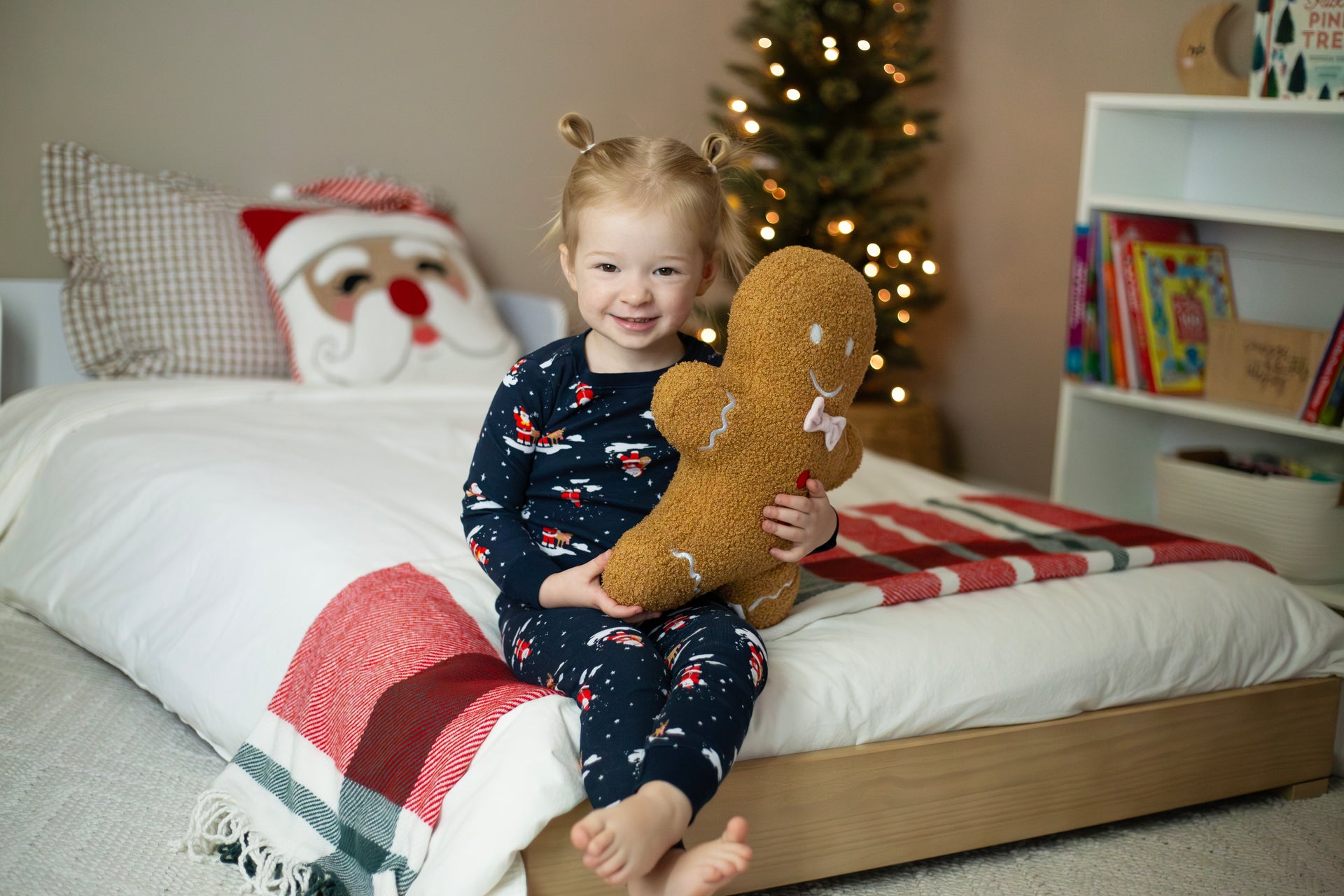 Child in festive pajamas holding a gingerbread plush toy in a cozy Christmas-themed bedroom.