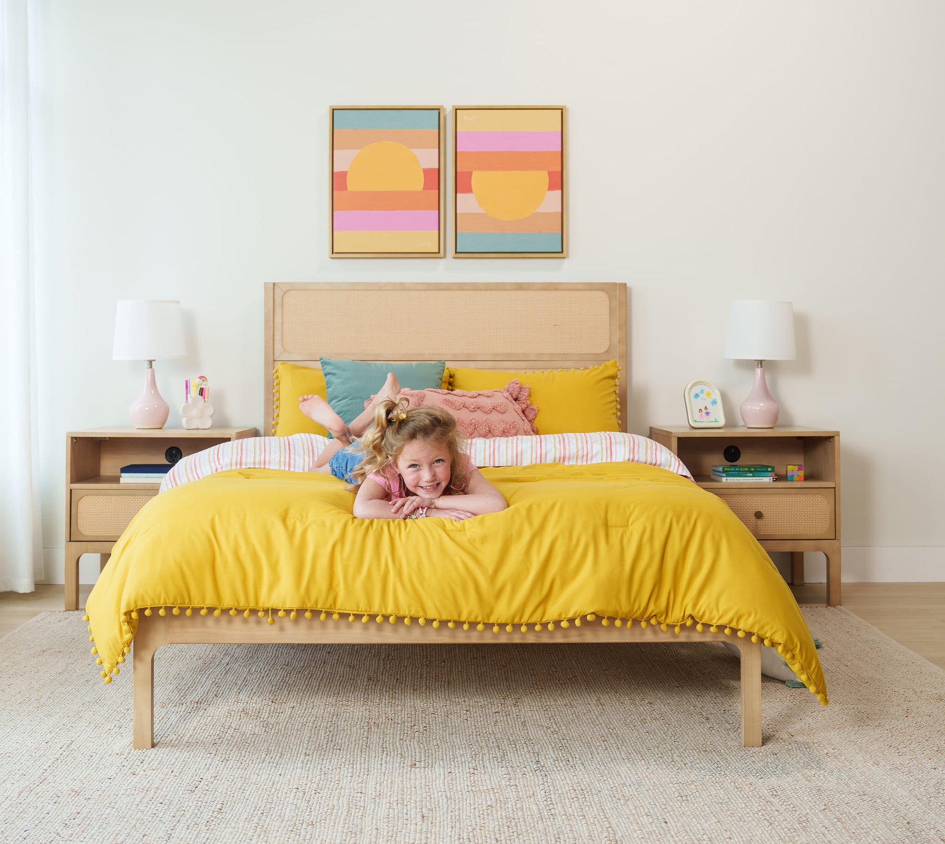 Children's bedroom with bright yellow bedding, woven rattan headboard, bedside lamps and colorful abstract wall art, smiling child lying on bed.