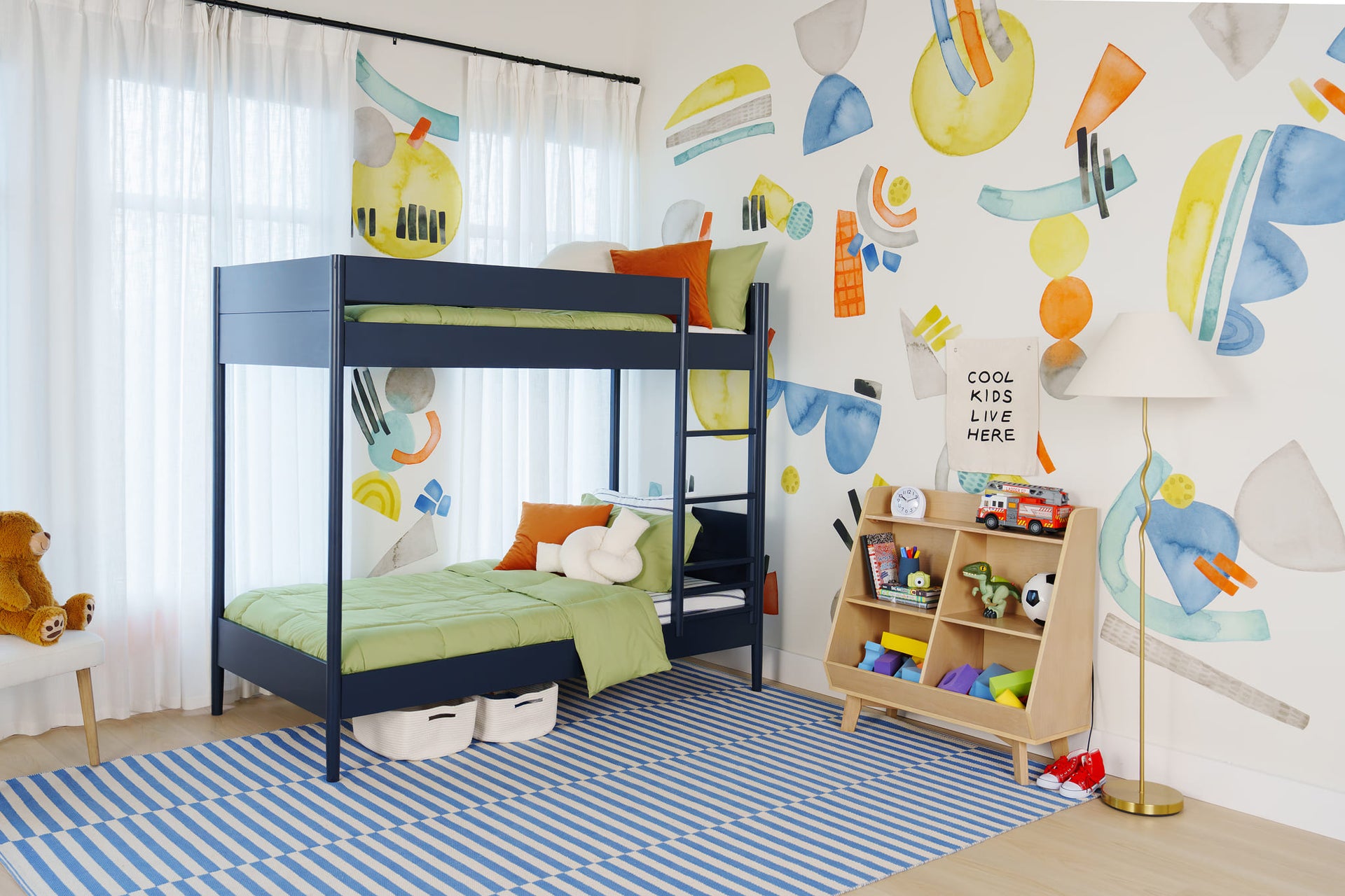 Bright modern kids' bedroom with navy bunk bed and green bedding, abstract colorful wall mural, wooden toy shelf with toys and blue-striped rug.
