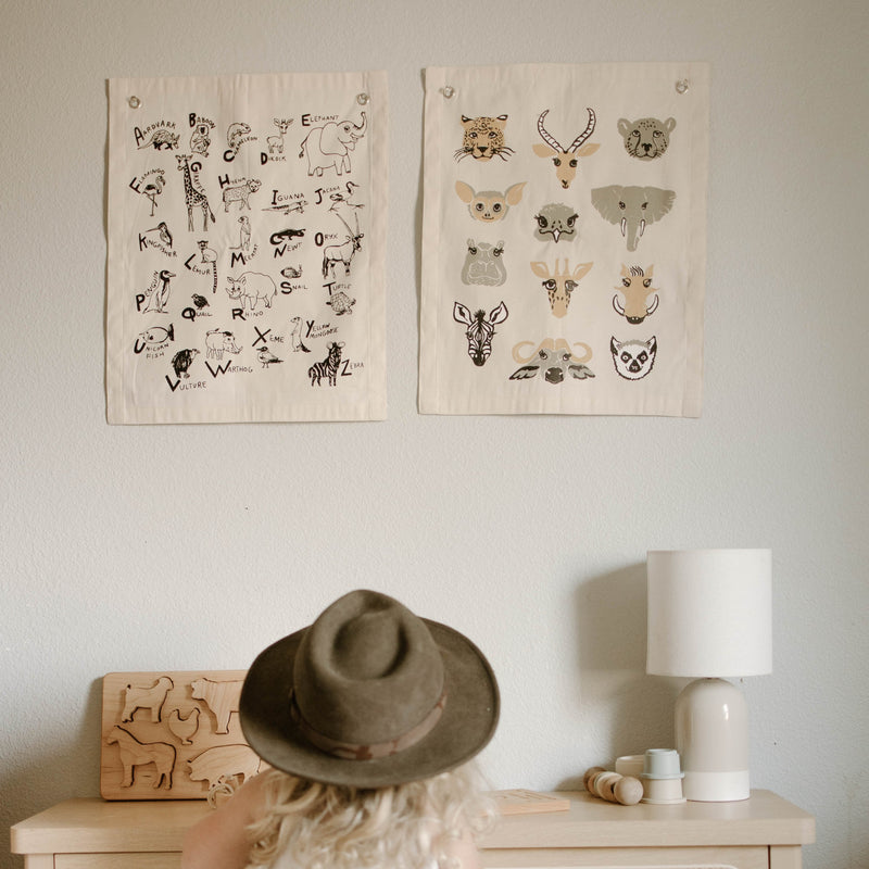 Child with hat looking at alphabet animal posters in a nursery room, featuring wooden animal puzzle pieces on a table.