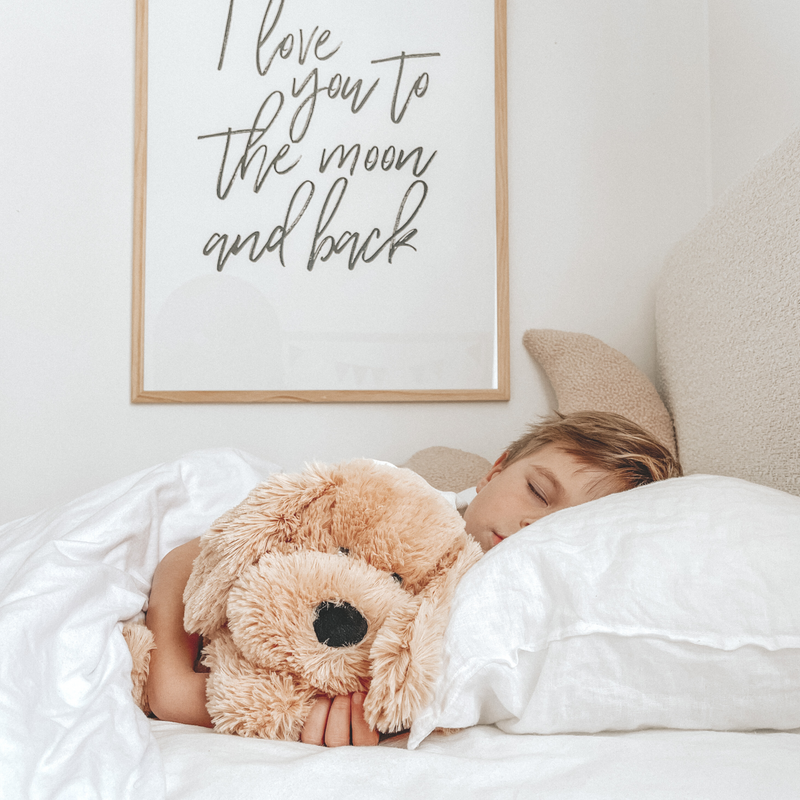 Child sleeping peacefully in bed hugging a teddy bear with "I love you to the moon and back" wall art, cozy bedroom atmosphere.