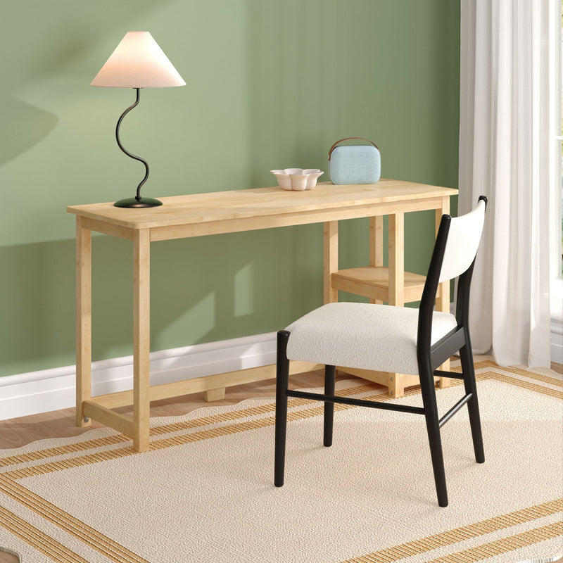 Minimalist Scandinavian home office desk with light wood table, curvy black table lamp, blue portable Bluetooth speaker, white ceramic bowl, upholstered chair, and sage green accent wall with sheer curtains.