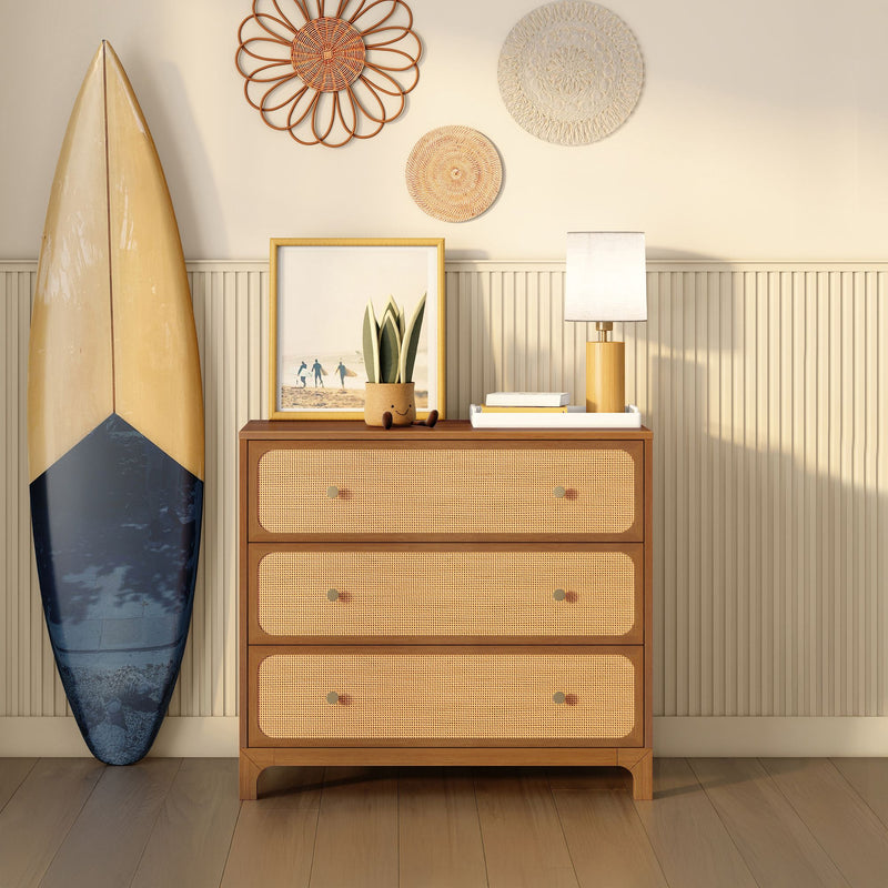Coastal boho entryway with woven rattan three-drawer dresser, leaning surfboard, table lamp, potted succulent, framed beach print and decorative wall baskets