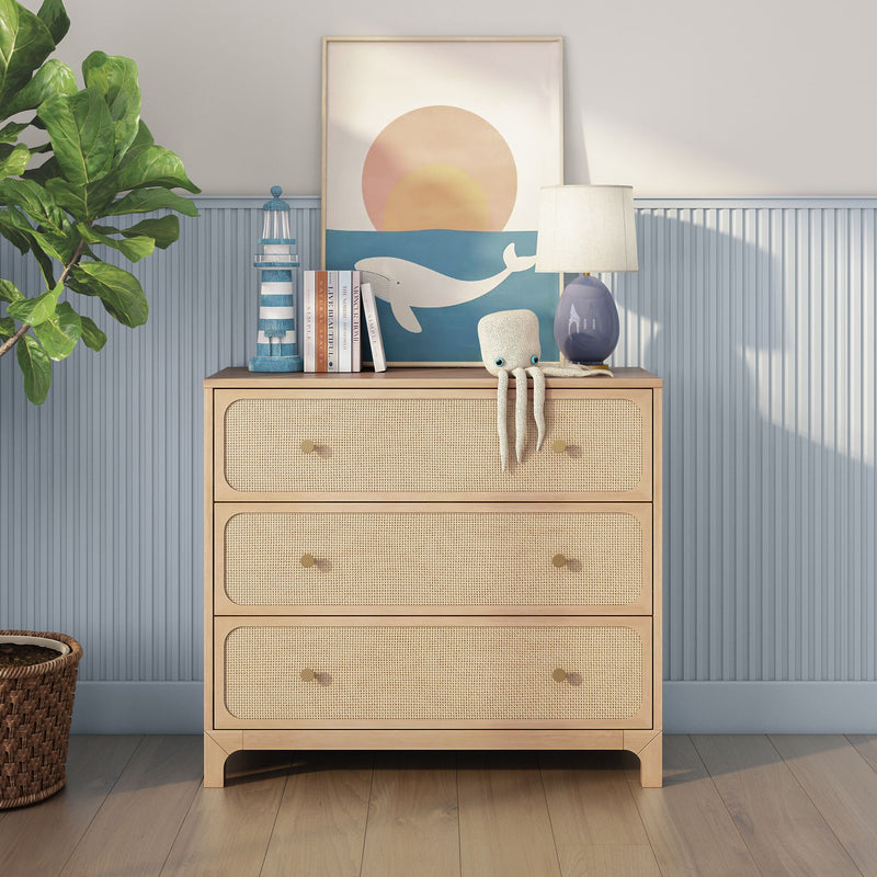 Light rattan three-drawer dresser in coastal nursery with whale sunset print, octopus plush toy, blue table lamp and potted fiddle-leaf fig