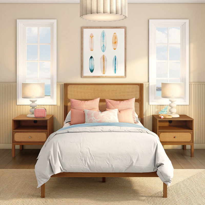 Coastal bedroom interior with rattan headboard, white bedding and pastel pillows, framed surfboard wall art centered between two tall windows with ocean view, matching wooden nightstands and table lamps for beachy modern decor