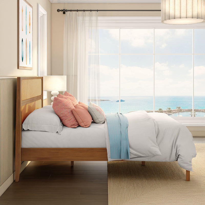 Light-filled coastal bedroom with wooden bed frame, layered pastel pillows, sheer curtains and large window overlooking turquoise ocean view