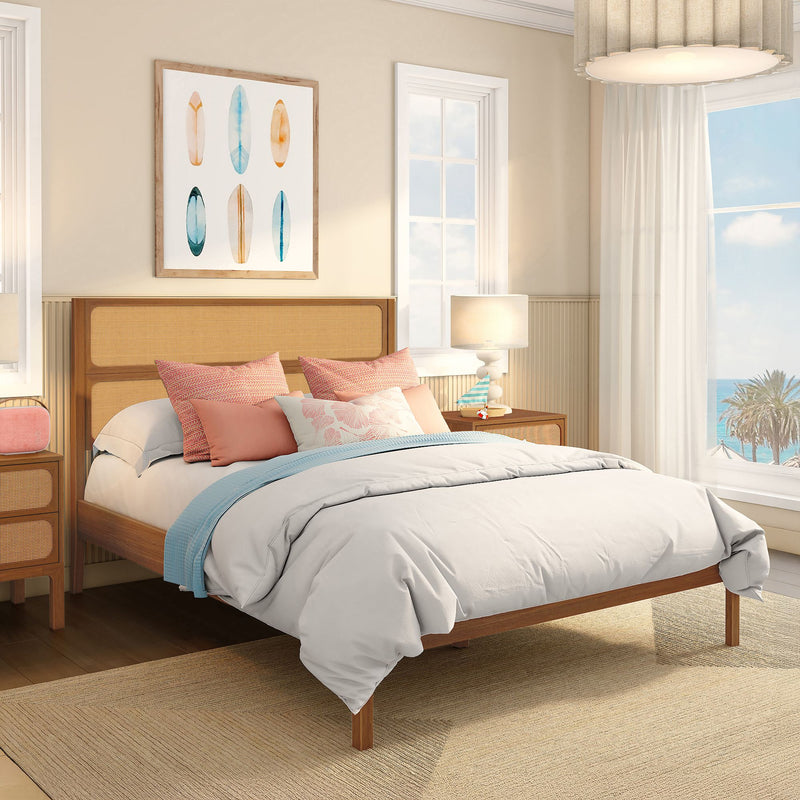 Coastal bedroom with rattan headboard bed, white comforter, coral and blue accent pillows, surfboard wall art, bedside lamp and ocean-view window