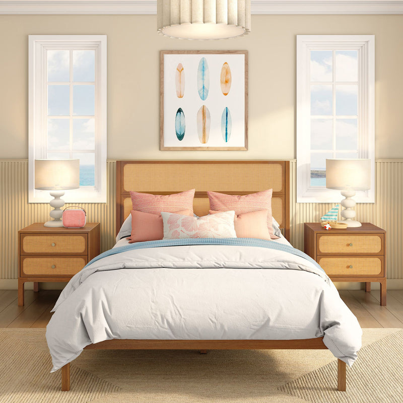 Beach-inspired bedroom with rattan headboard, pink pillows and white duvet, surfboard wall art, wooden nightstands, table lamps and ocean-view windows.