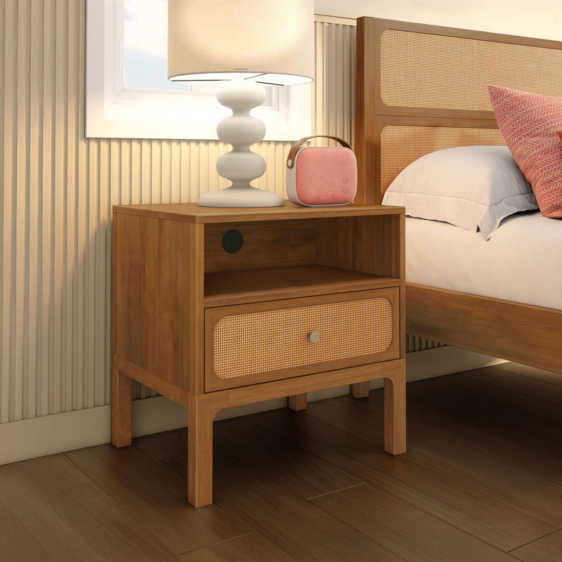 Mid-century modern wooden nightstand with woven rattan drawer, white table lamp and pink portable Bluetooth speaker on top beside an upholstered bed in a warm oak bedroom interior with hardwood floors