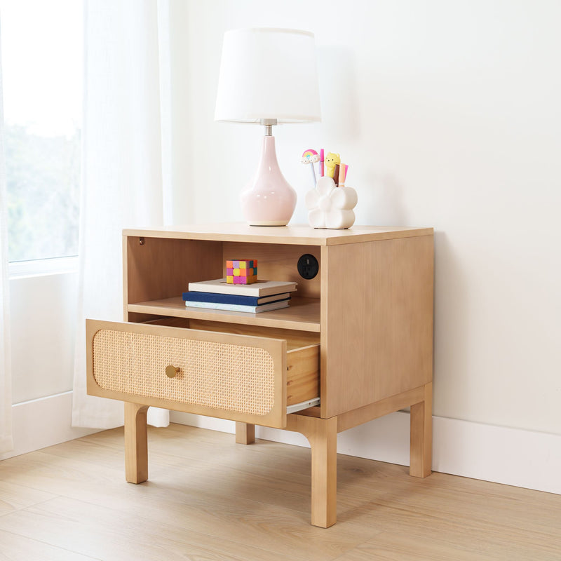 Light wood nightstand with rattan drawer, pink ceramic table lamp, white flower pen holder with colorful pens and stacked books — minimalist kids' room decor by sunlit window