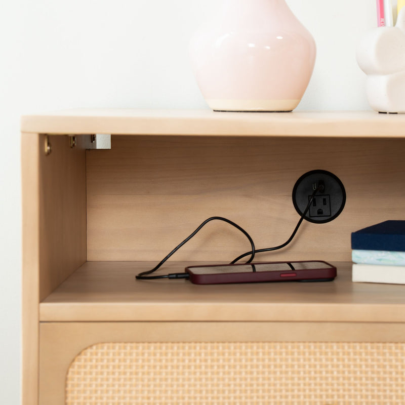 Wooden bedside shelf with built-in power outlet and charging smartphone, pink ceramic vase, stacked books and rattan drawer front