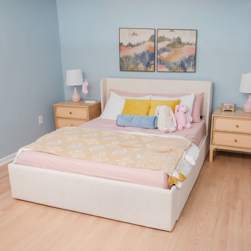 Cozy pastel bedroom with upholstered bed, pink bedding, yellow and blue accent pillows, plush bunny toys, wooden nightstands and abstract wall art