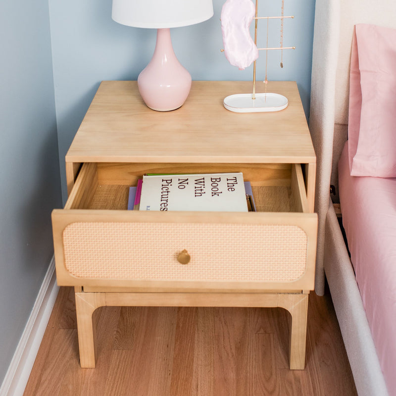 Light wood mid-century nightstand with open drawer showing "The Book With No Pictures", pink ceramic lamp, gold jewelry stand and pastel pink bedding on hardwood floor