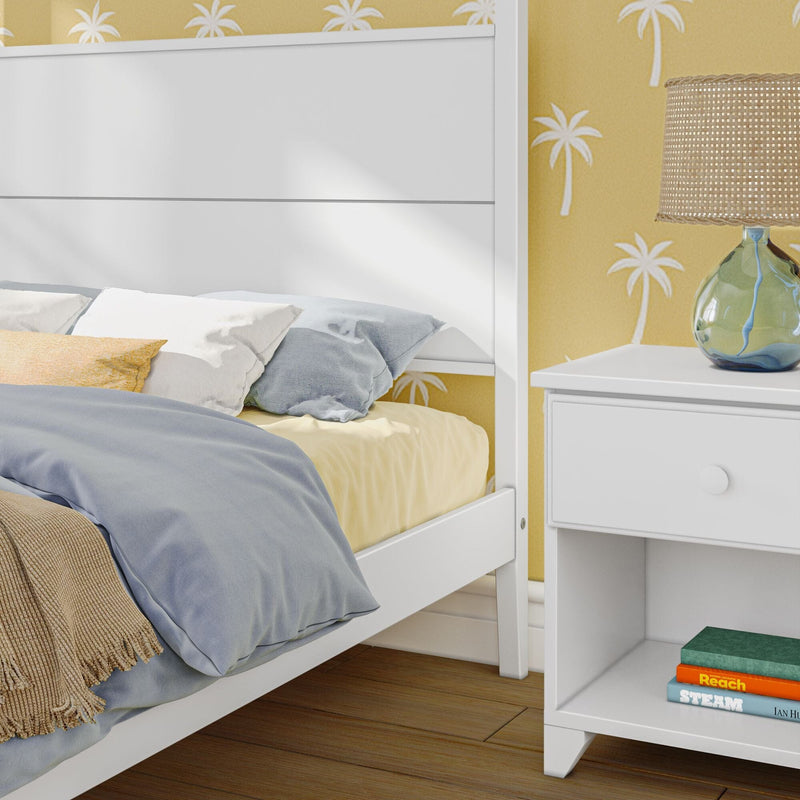 Close-up of coastal bedroom interior with white wooden bed frame, blue and yellow bedding, palm-print yellow wallpaper, rattan lamp on a white nightstand with stacked books