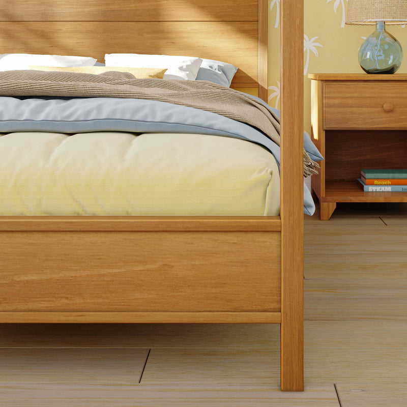 Close-up of a wooden bed frame with yellow bedding, blue pillows and textured beige throw beside a wooden nightstand with glass lamp and palm-print wallpaper