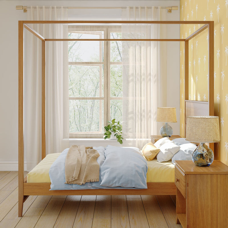 Cozy modern bedroom with wooden four‑poster canopy bed, soft blue bedding, yellow palm‑print accent wall, sheer curtains, potted plant and woven bedside lamp