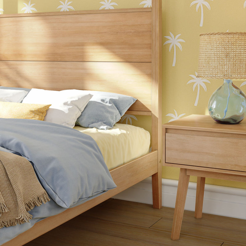 Coastal bedroom close-up of a light wood bed frame and nightstand with woven rattan lamp, soft blue and yellow bedding, and palm-print yellow wallpaper