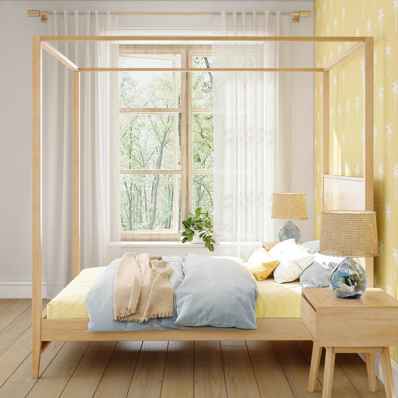 Scandinavian-style bedroom with light wood four-poster canopy bed, pastel blue and yellow bedding, sheer curtains, palm-print yellow accent wall and rattan bedside lamp