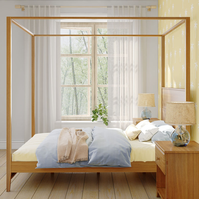 Modern wooden canopy bed in a sunlit Scandinavian bedroom with pastel blue and yellow bedding, woven bedside lamps on wooden nightstands, potted plant by the window, natural wood floors and palm-pattern yellow accent wall, minimalist cozy interior