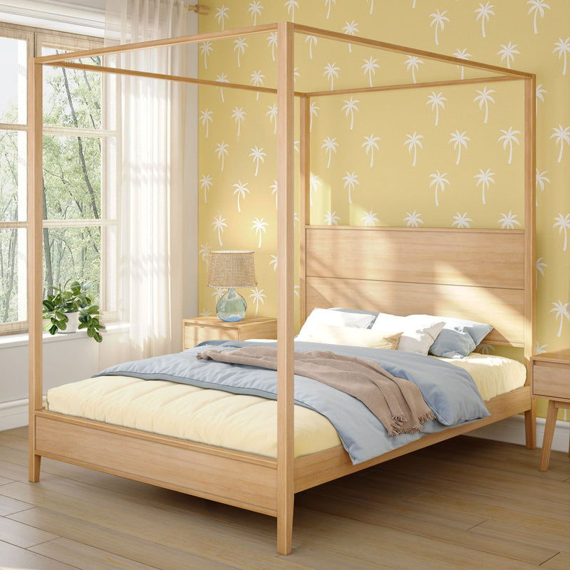 Sunlit modern coastal bedroom with natural wood four-poster canopy bed, blue and cream bedding, yellow palm-print wallpaper, bedside lamp and potted plant