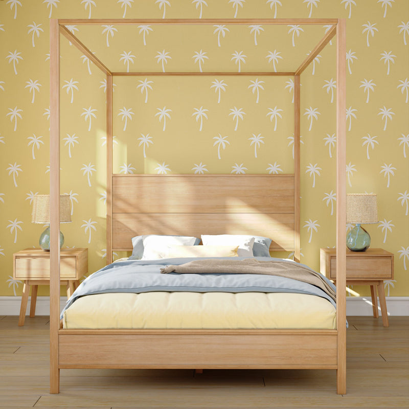 Sunlit wooden four-poster bed with blue and yellow bedding against palm-print yellow wallpaper, flanked by matching wooden nightstands and glass lamps — coastal bedroom interior