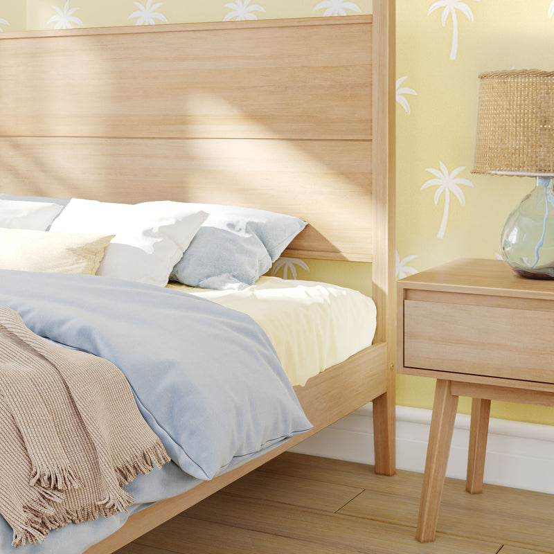 Modern coastal bedroom with light wood bed and nightstand, blue duvet and pillows, beige knit throw, yellow palm-print wallpaper and rattan table lamp