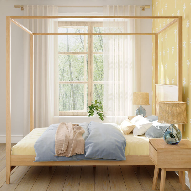 Sunlit Scandinavian bedroom with natural wood canopy bed, blue bedding, rattan bedside lamps and yellow palm-print accent wall