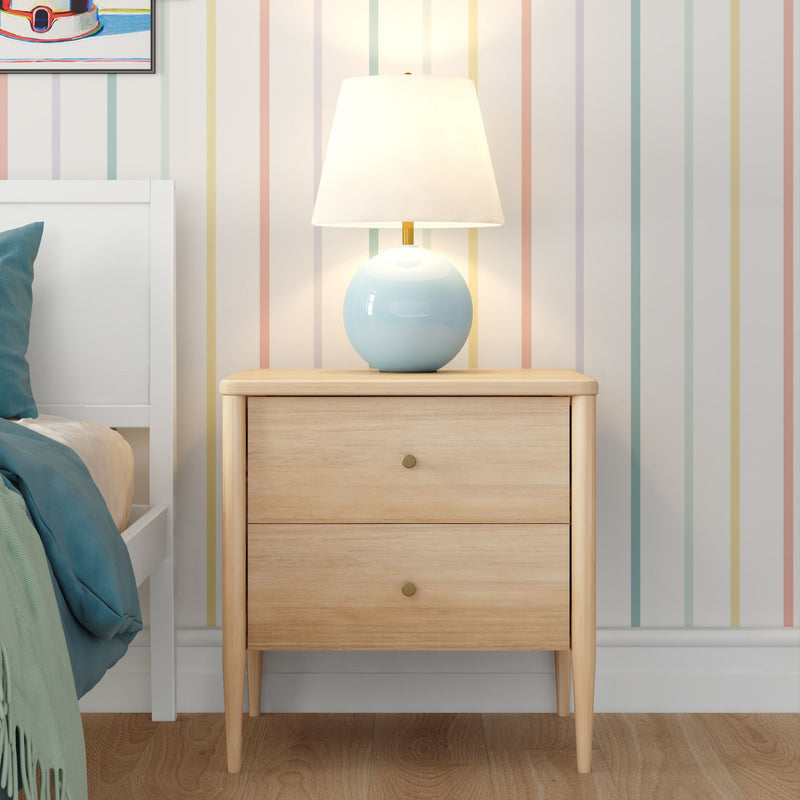 Wooden two-drawer bedside table with tapered legs and blue ceramic table lamp beside a white bed with teal bedding and pastel striped wallpaper — modern cozy bedroom decor