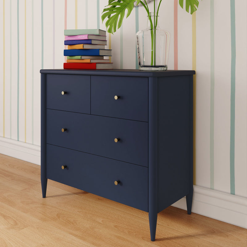 Navy blue dresser with gold knobs topped by a stack of colorful books and a glass vase holding monstera leaves against pastel striped wallpaper on a hardwood floor