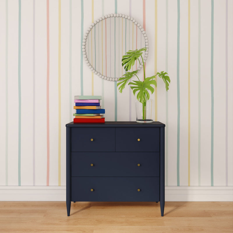 Navy dresser with brass knobs, stacked colorful books and glass vase holding a Monstera plant under a round beaded mirror on pastel vertical striped wallpaper