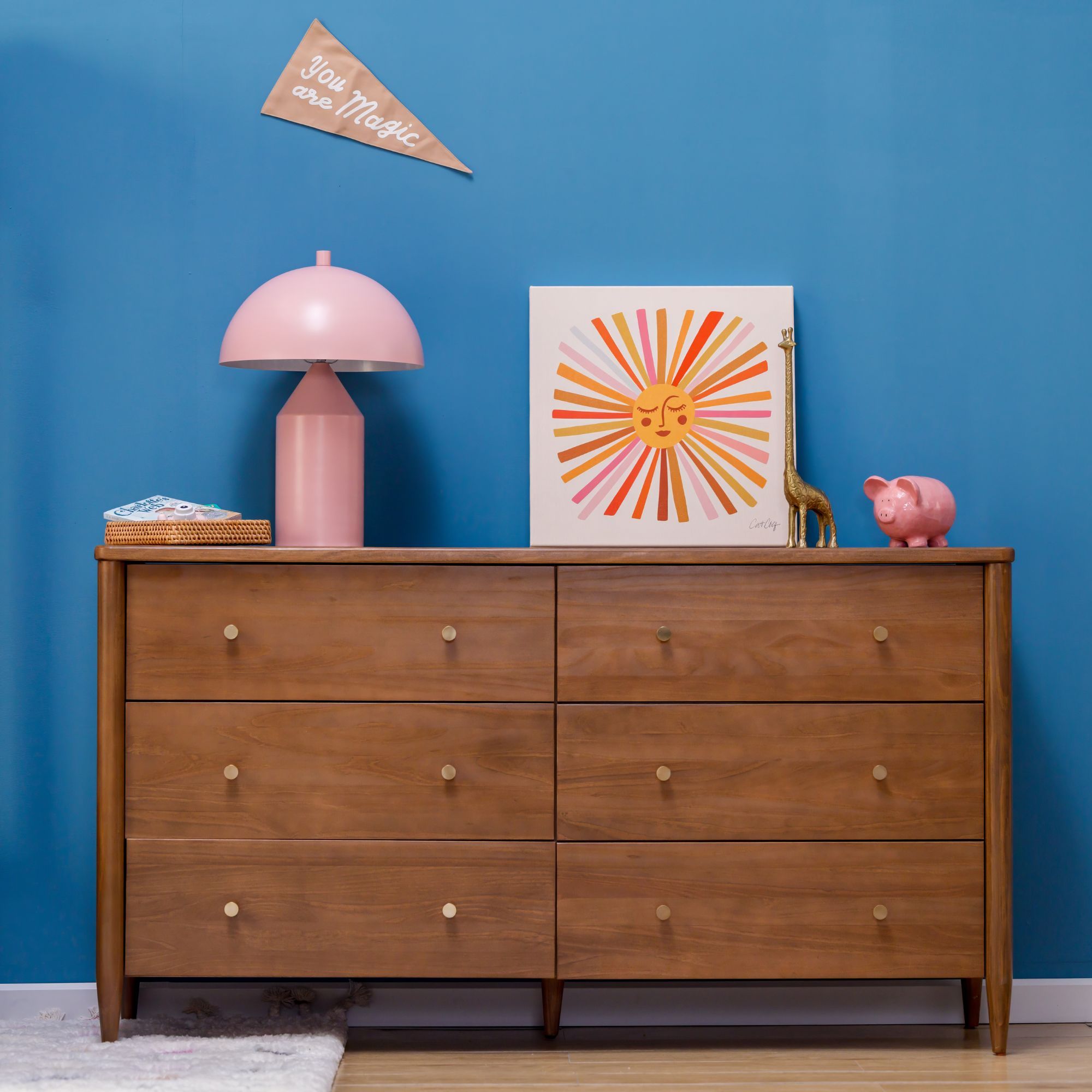 A warm wood modern dresser with 6 drawers in a blue room. There is a pink lamp, piggy bank, and sun art on the dresser.